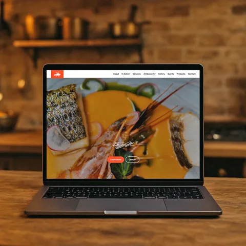 Website preview of cbdcuisine.com displayed on a laptop placed on a rustic chef’s kitchen table, showing Chef Ben’s dish with prawns and golden sauce in the hero background.